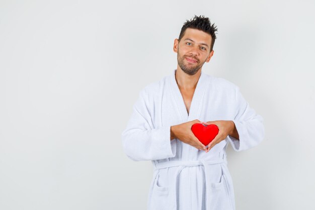 Young man holding red heart in white bathrobe and looking joyful. front view.