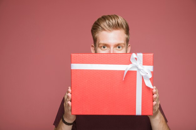 Young man holding red present box in front of his mouth against colored background