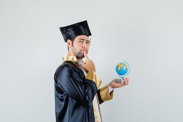 Young man holding school globe in graduate uniform and looking thoughtful .