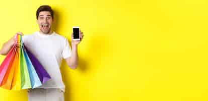 Free photo young man holding shopping bags and showing mobile phone screen money application standing over yell