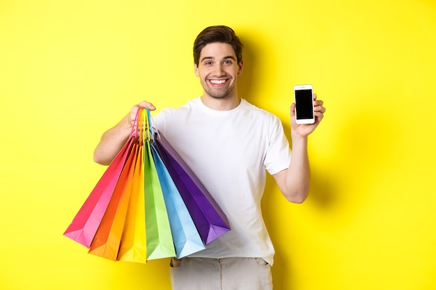 노란색 배경 위에 서 있는 젊은 남자가 쇼핑백을 들고 휴대폰 화면, 화폐 응용 프로그램을 보여줍니다.