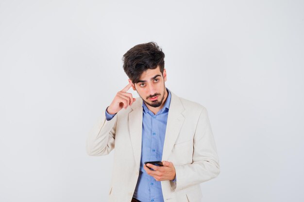 Young man holding smartphone and putting index finger on ear in blue t-shirt and white suit jacket and looking serious