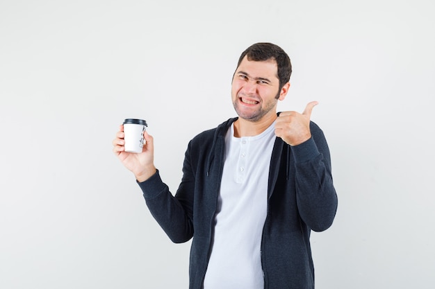 Young man holding takeaway coffee cup and showing thumb up in white t-shirt and zip-front black hoodie and looking happy. front view.