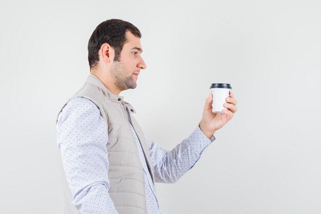 Young man holding takeaway cup of coffee and looking at it in beige jacket and looking serious , front view.