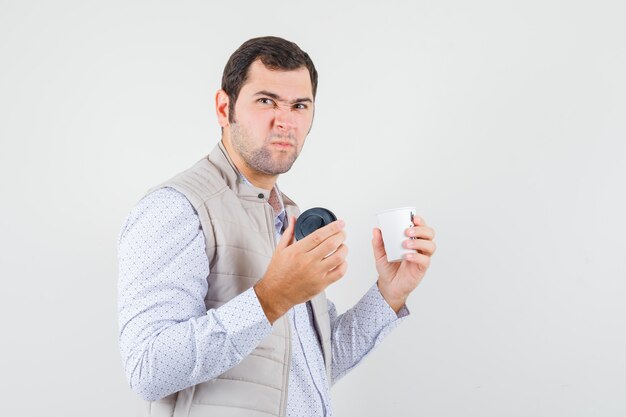 Young man holding takeaway cup of coffee and opening it in beige jacket and looking displeased , front view.