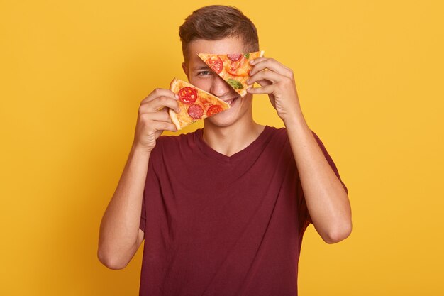 若い男が2つのおいしいピザを手に持って、おいしい製品で彼の目を覆っています。