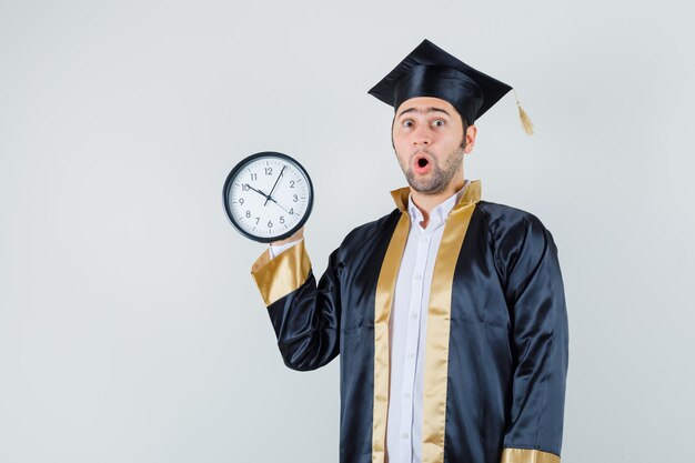 Young man holding wall clock in graduate uniform and looking surprised , front view.