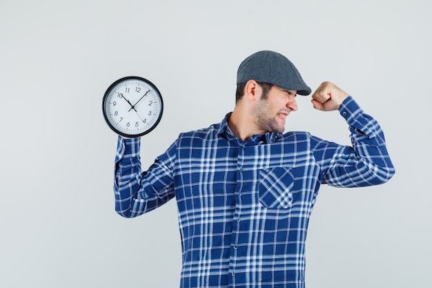 Free photo young man holding wall clock in shirt, cap and looking blissful. front view.