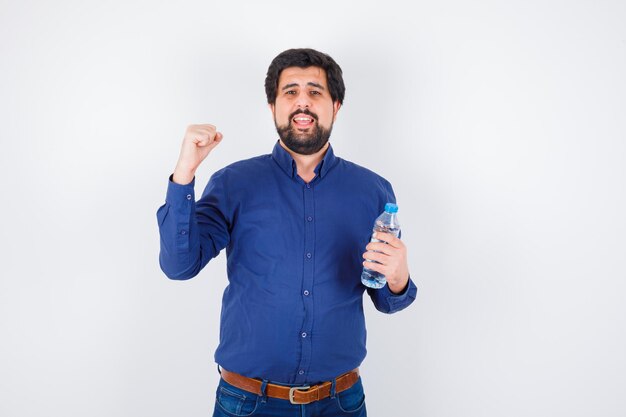 Young man holding water bottle and showing power gesture in blue shirt and jeans and looking optimistic , front view.