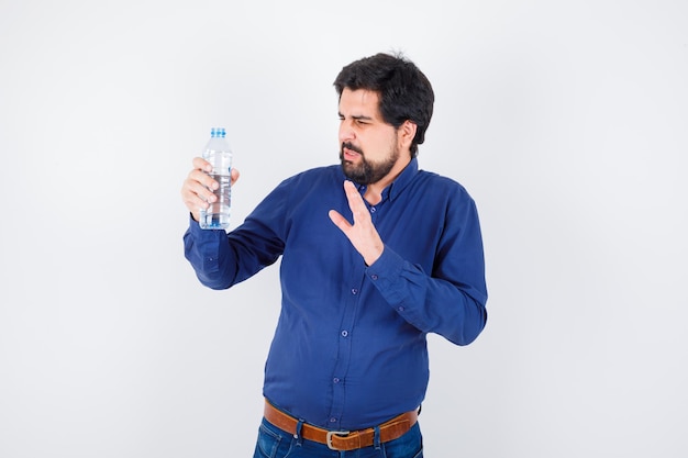 Молодой человек держит бутылку с водой и протягивает к ней руку в синей рубашке и джинсах и выглядит недовольным. передний план.