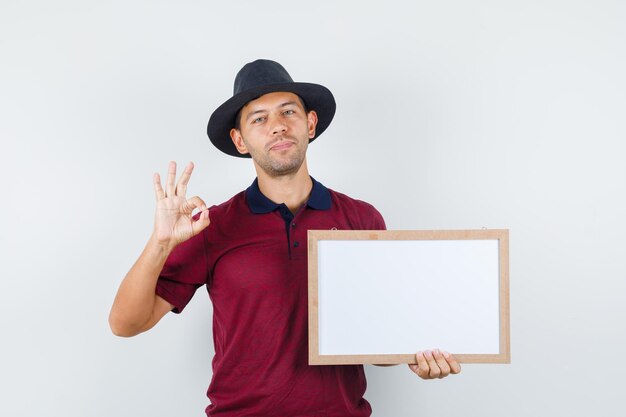 Young man holding white board with ok sign in t-shirt, hat and looking pleased. front view.