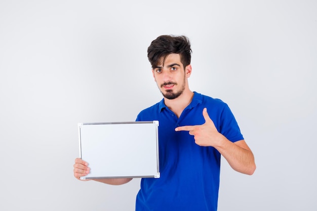 Young man holding whiteboard and pointing to it in blue t-shirt and looking serious
