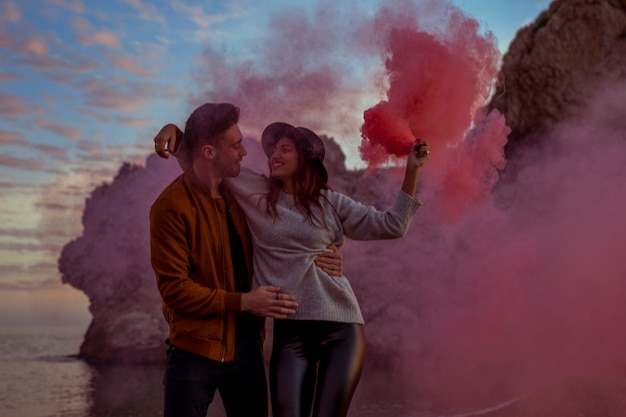 Young man holding woman with pink smoke bomb on sea shore