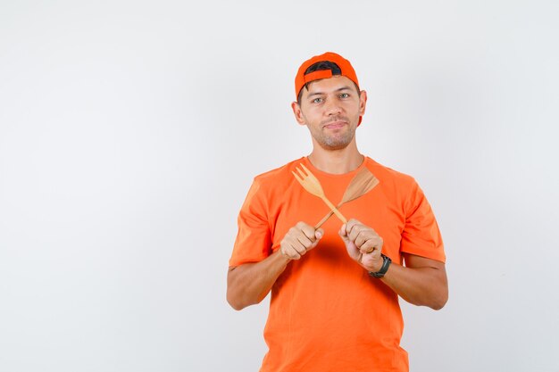 오렌지 티셔츠와 모자에 나무 포크와 주걱을 들고 자신감을 찾고 젊은 남자