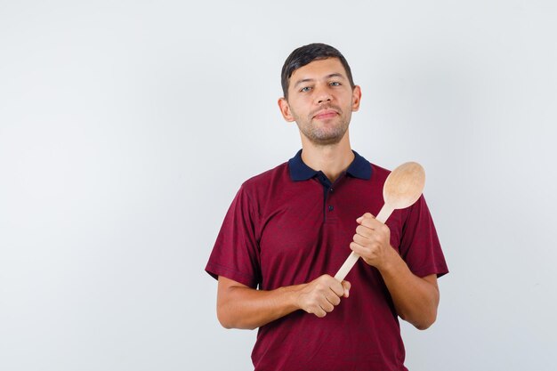 Young man holding wooden spoon in t-shirt and looking cheerful. front view.