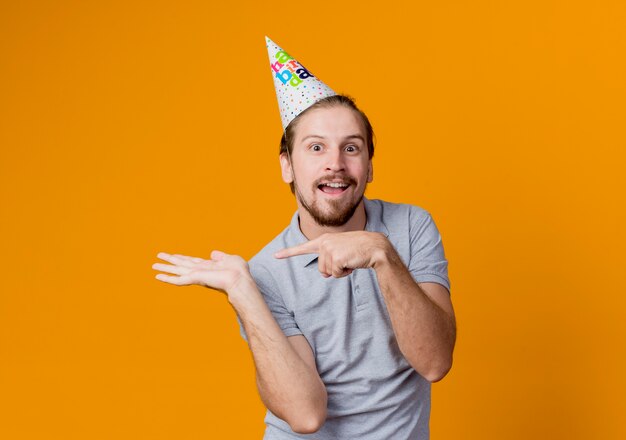 オレンジ色の壁の上に立っている側の誕生日パーティーのコンセプトを腕で提示し、指で指しているホリデーキャップの若い男