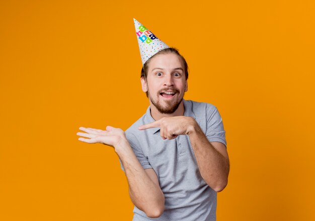 Young man in holiday cap  presenting with arm and pointing with finger to the side birthday party concept standing over orange wall