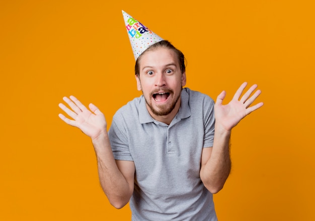 Young man in holiday cap surprised and confused smiling birthday party concept standing over orange wall