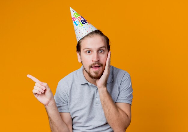 young man in holiday cap  surprised pointign with finger to the side birthday party concept standing over orange wall