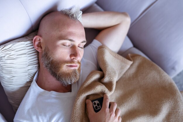 自宅の若い男が携帯電話でソファで眠りに落ちた