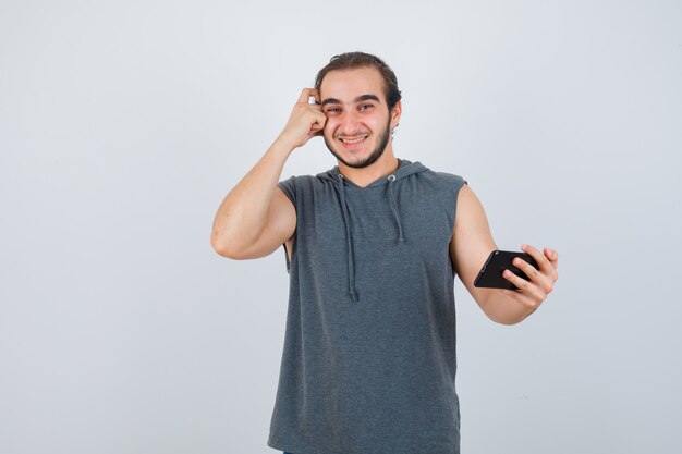 Молодой человек в футболке с капюшоном держит телефон в руке, держит руку на голове и выглядит веселым, вид спереди.