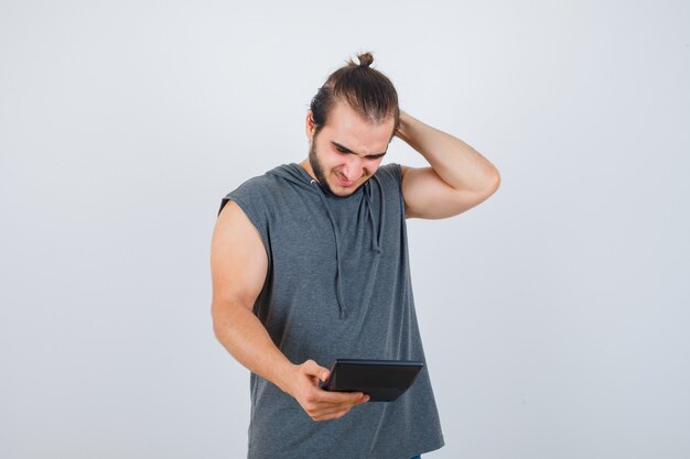 Young man in hoodie holding hand behind head, looking at calculator and looking pleased , front view.