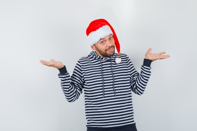 Young man in hoodie, Santa Claus hat showing helpless gesture and looking indecisive , front view.