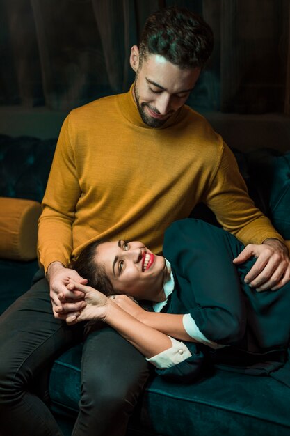 Young man hugging cheerful woman lying on legs on settee