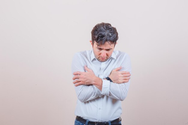 young man hugging himself in white shirt, jeans and looking anxious