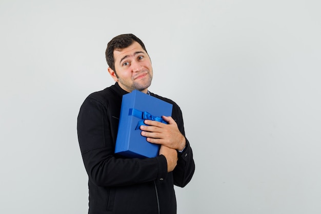 Young man hugging present box in shirt, jacket and looking cute. 