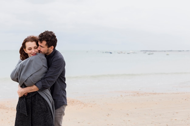 Young man hugging woman in grey blanket on sea shore