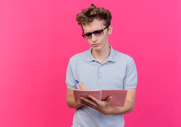 Бесплатное фото Молодой человек в черных очках в серой рубашке поло держит блокнот с ручкой и смотрит на него с серьезным хмурым лицом поверх розового