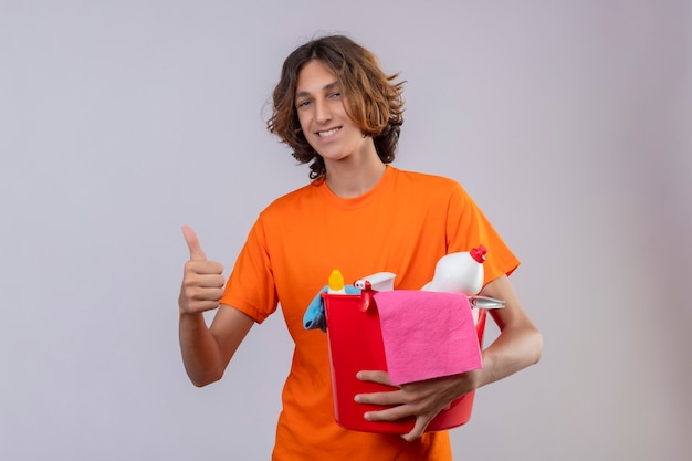 Бесплатное фото Молодой человек в оранжевой футболке держит ведро с инструментами для уборки, глядя в камеру, весело улыбаясь, показывая пальцы вверх, стоя на белом фоне