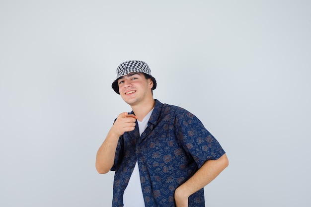 Молодой человек в белой футболке, рубашке с цветочным рисунком, кепке, указывающей на камеру указательным пальцем и счастливой