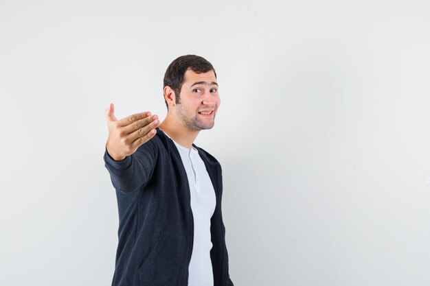 Young man inviting to come in white t-shirt and zip-front black hoodie and looking optimistic , front view.