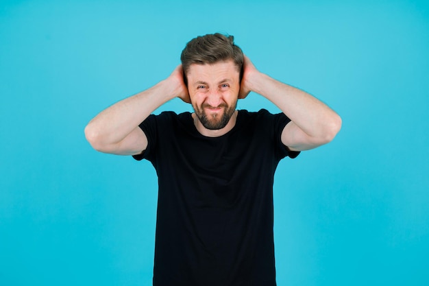 Young man is covering his ears with hands on blue background