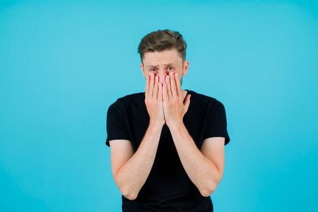 Young man is looking at camera by covering cheeks and mouth with hands on blue background