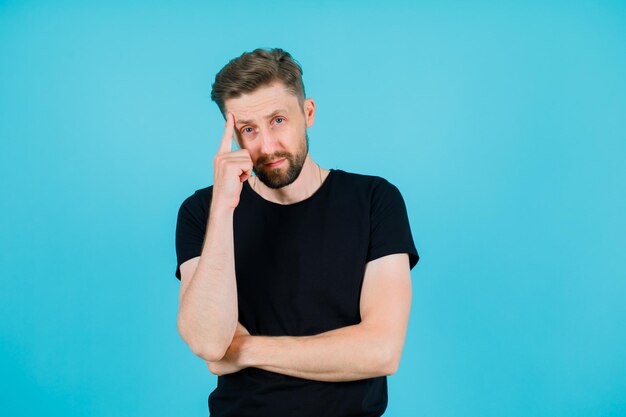 Young man is looking at camera by holding forefinger on temple on blue background