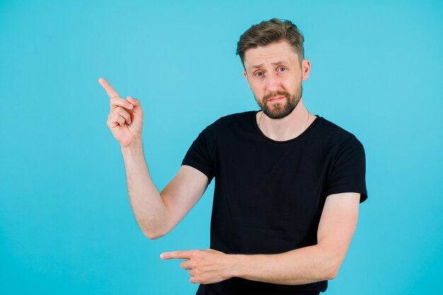 Young man is looking at camera by pointing up with forefinger on blue background