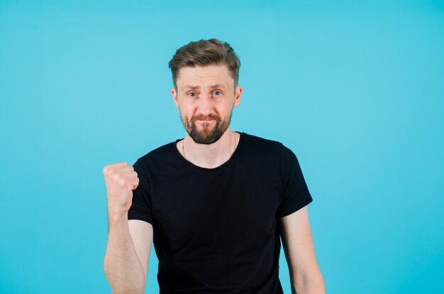 Young man is looking at camera by raising up his fist on blue background