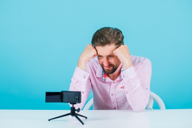 Young man is looking at his mini camera by holding fists on forehead on blue background