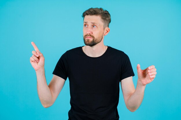 Young man is looking up an dpointing left up with forefinger on blue background