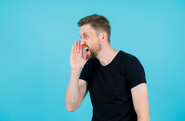 Young man is screaming by holding hand near mouth on blue background