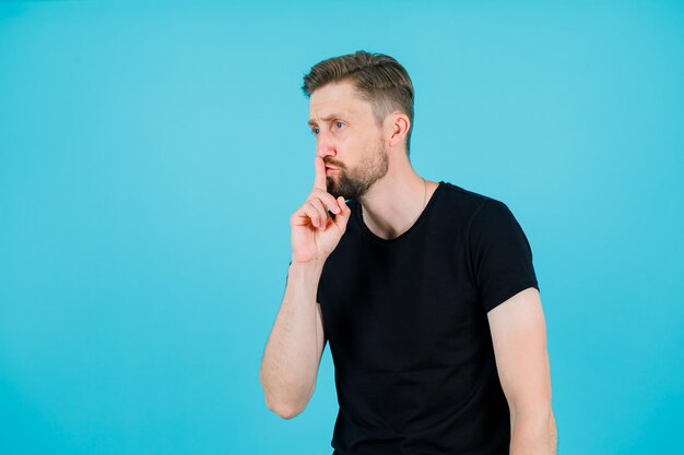 Young man is showing silence gesture by holding forefinger on lips on blue background