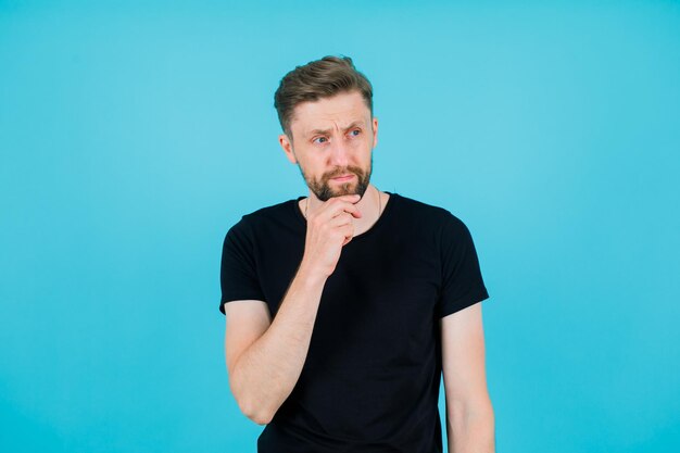 Young man is thinking by holding hand on chin on blue background