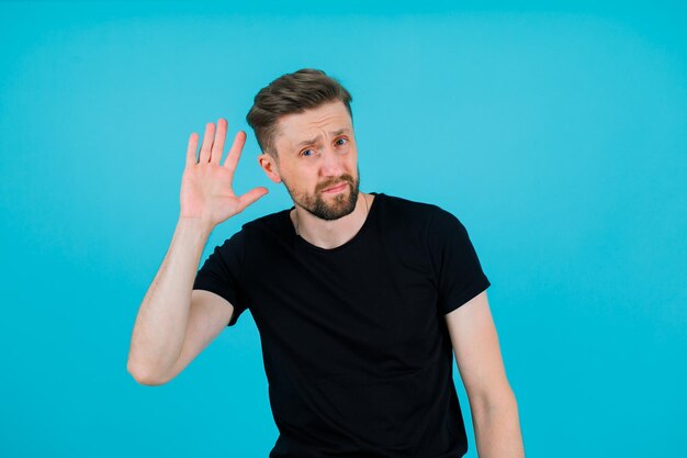 Young man is trying to hear by holding hand near ear on blue background