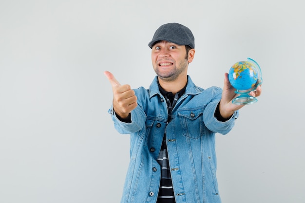 Young man in jacket,cap holding globe while showing thumb up and looking glad