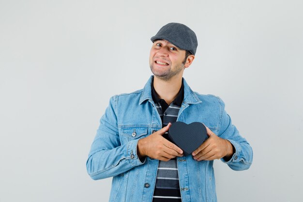 Young man in jacket,cap holding heart-shaped box and looking pleased