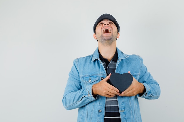 Young man in jacket,cap holding heart-shaped box while looking up and looking merry