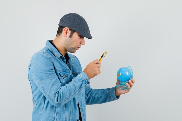 Young man in jacket,cap looking mini globe with magnifier and looking careful .